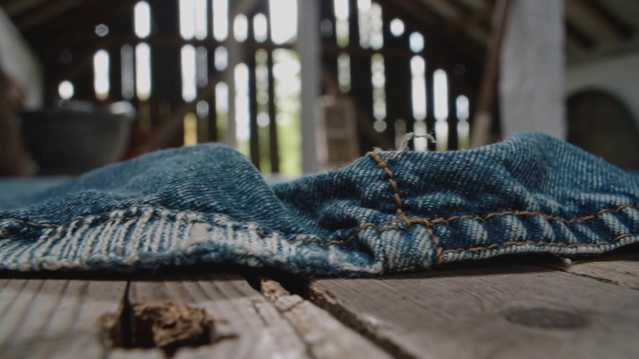 Pushing camera bringing worn blue denim hem into focus on barn floor, showing frayed stitching