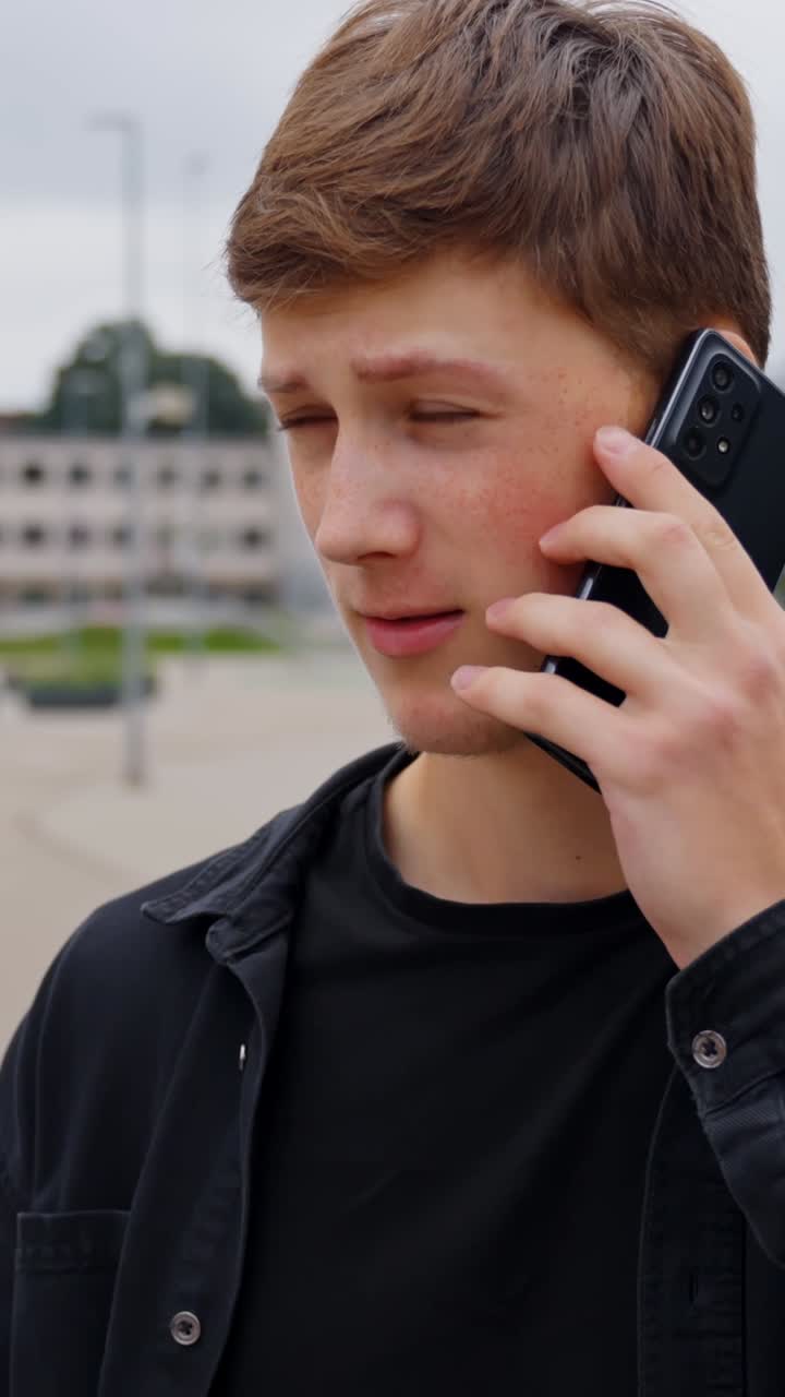 A young man with brown hair wearing a black jacket is making a phone call while walking outdoors in a modern urban city park, surrounded by plants, blurred architecture, vertical shot in natural light