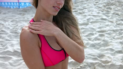 una mujer con un bikini rosa y negro, aplicándose crema solar en el hombro en la playa, con el pelo ondeando al viento