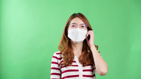 happy young asian woman with eyeglasses taking off medical mask, looking at camera amd smiling when corona virus pandemic is over, green screen background