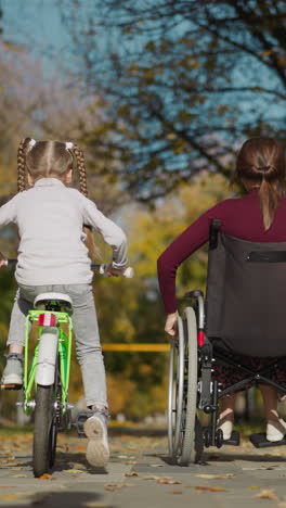 mother with daughter and son spends free time in autumn park. woman in wheelchair moves by spinning big wheels with hands. girl and boy ride bicycles backside view