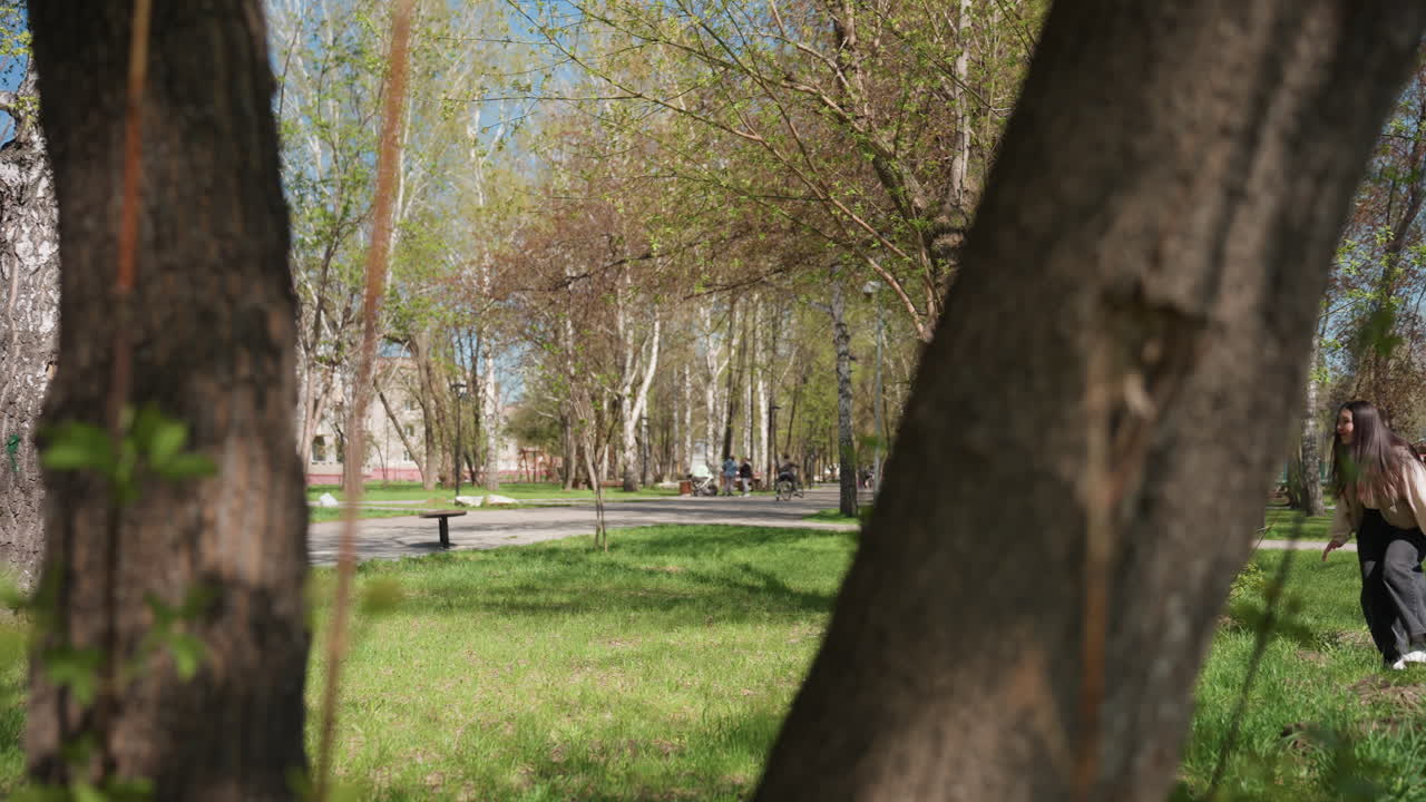 niños jugando al escondite cerca de un árbol, parque soleado de primavera con troncos de abedul, hierba verde, dos jóvenes persiguiéndose alrededor de un tronco nudoso, bancos dispersos y edificios distantes, chaquetas informales, movimiento lúdico
