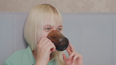 blonde woman with bangs wearing green shirt smiles warmly while sipping from brown glass cup, she appears joyful and relaxed in soft indoor lighting, seated against cushioned bed headboard