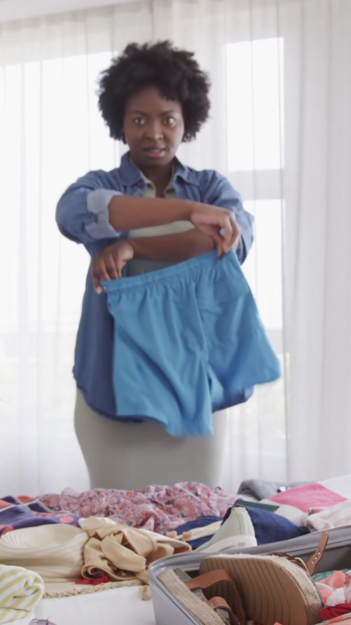 Vertical video: Female pals packing suitcase on bed pal holding sandals urging checklist checking