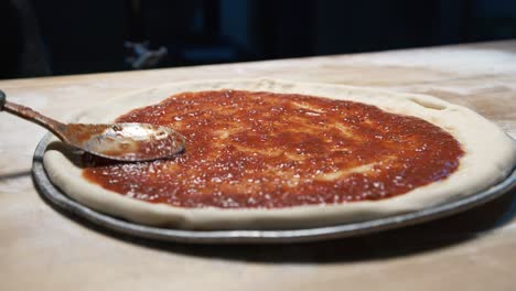 a close-up of a pizza dough on a steel tray on wooden counter while an expereinced chef finishing put juicy red sauce on it with a adicid