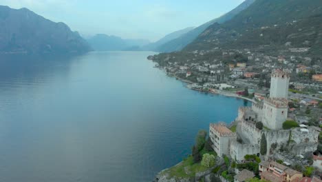 Aerial-view-of-Malcesine-Castle-tower-overlooking-Lake-Garda,-with-the-town,-rooftops,-and-mountains,-Italy