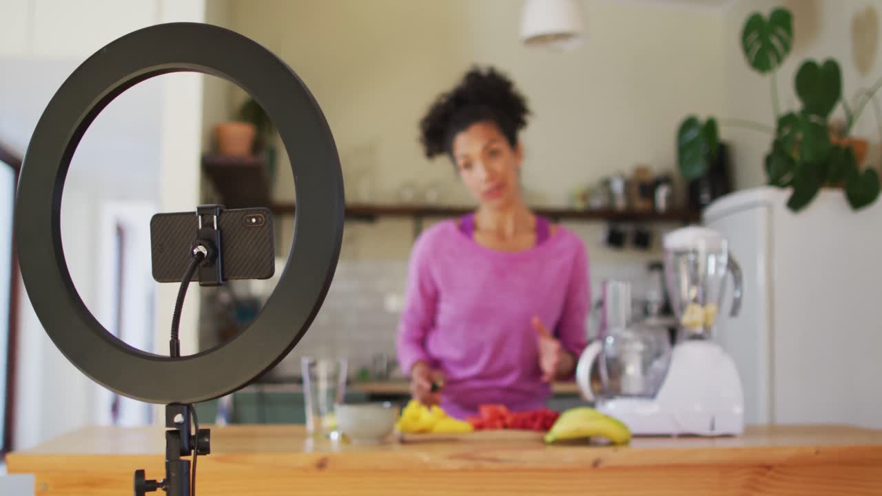 Vlogger donna di etnia mista che prepara succo di frutta e registra con lo smartphone in cucina a casa.