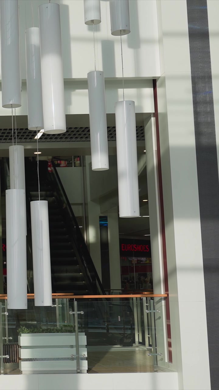 Close-up de duas pessoas caminhando juntas em um shopping center. O fundo apresenta luzes cilíndricas brancas de vidro penduradas. A luz do dia brilhante ilumina espaços interiores modernos.