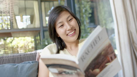 happy asian woman sitting on couch and reading book in sunny living room, slow motion