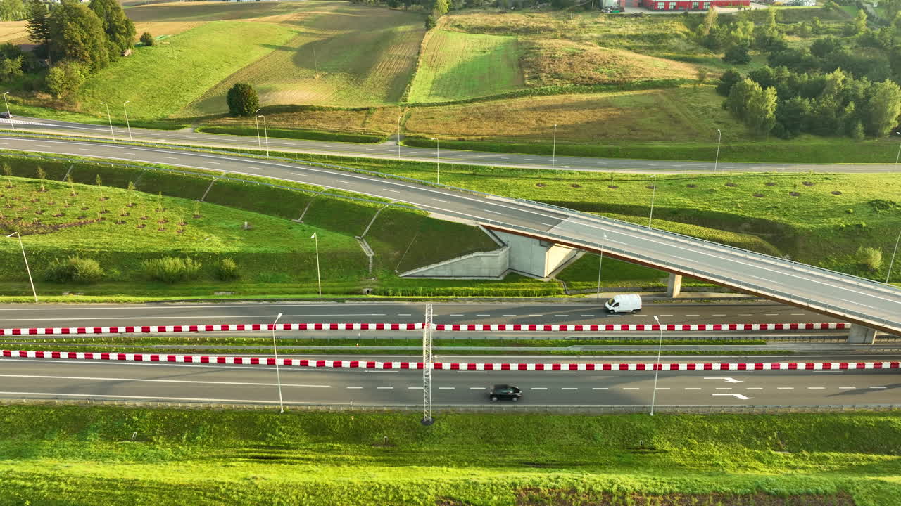 Aerial view of a modern highway overpass cutting through green rural hills and fields. A white van and black car drive past red-and-white safety barriers in bright sunlight