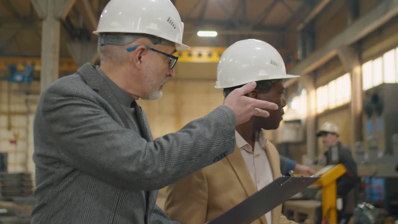 Multiethnic Engineers in Hardhats Walking and Speaking in Machinery Plant
