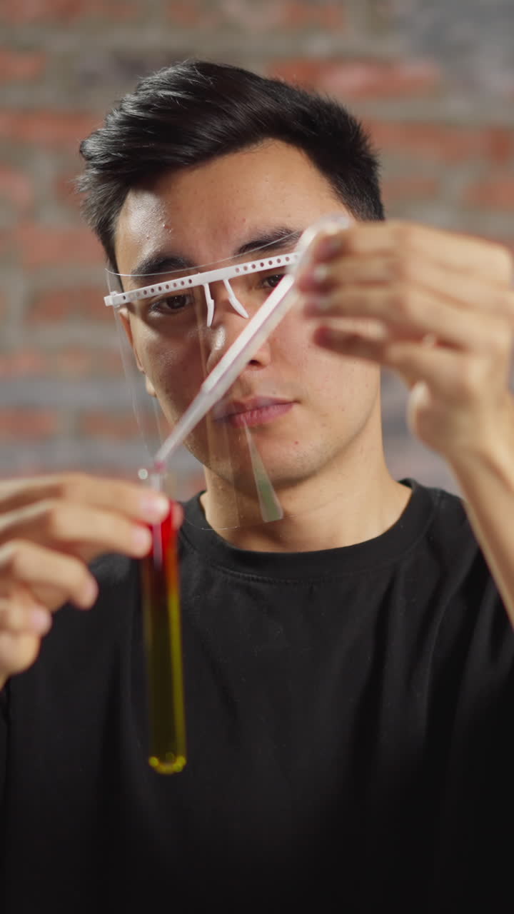 Young Asian male chemist with face shield adds reagent into tube with yellow liquid doing tests in scientific laboratory of university closeup