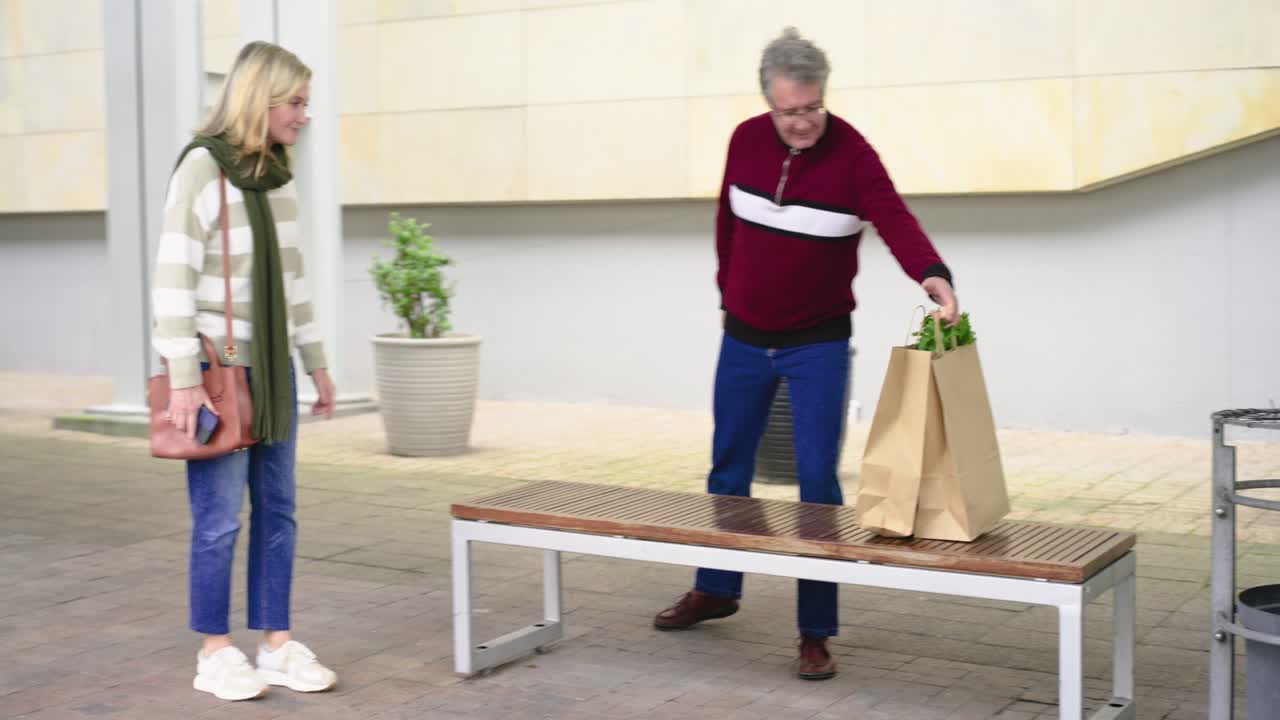 Couple walking in courtyard past metal screen placing grocery bags on bench and settling with smile