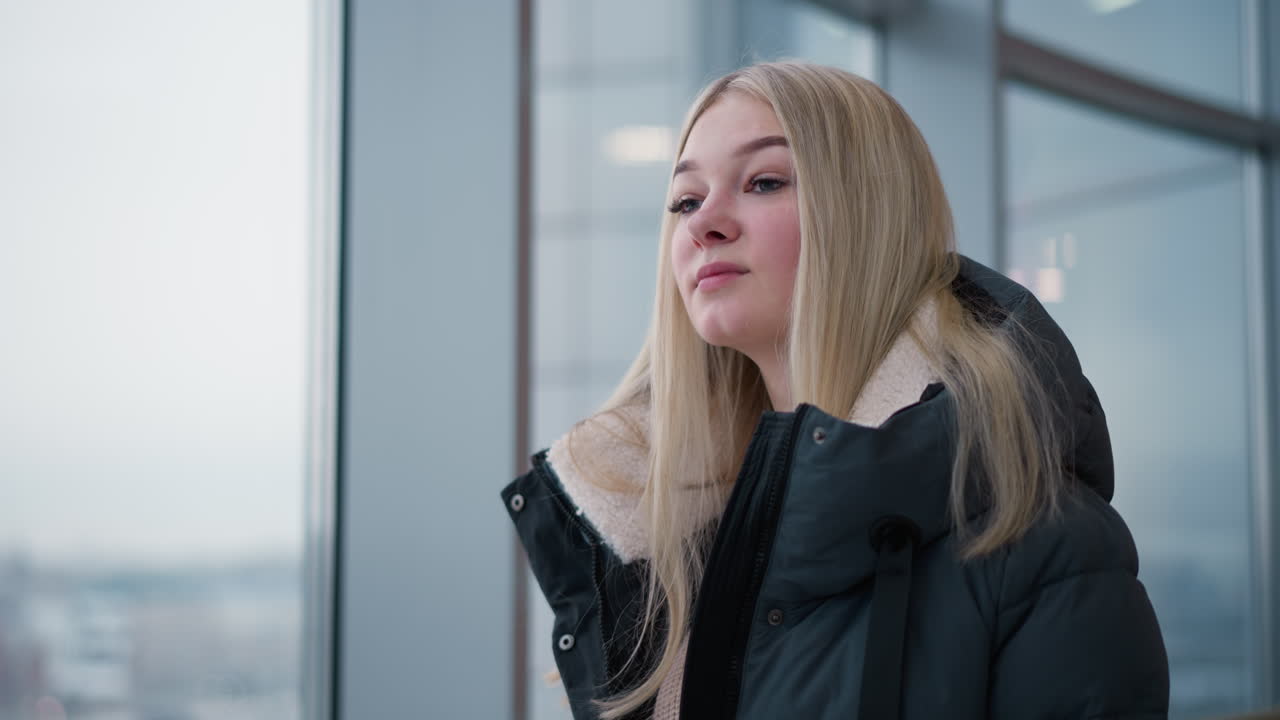 그녀의 어깨 아래로 머리카락을 캐스케이딩하는 여인이, 유리 창문을 통해 볼 수 있는 도시 배경과 함께 상하는 모습을 보이고, 평온과 반성의 순간