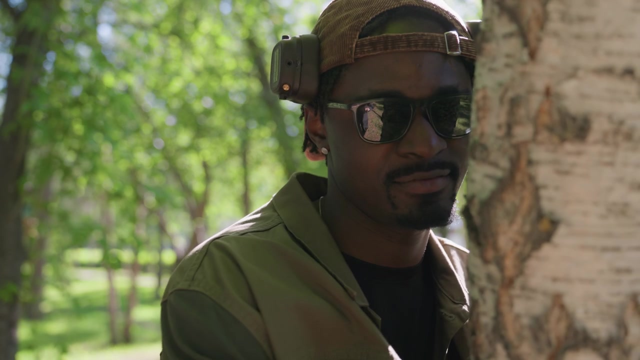 Man wearing cap and jacket touches tree bark in park with green trees sunlight filtering through leaves. Man examines texture of birch trunk while standing outdoors on grassy area surrounded
