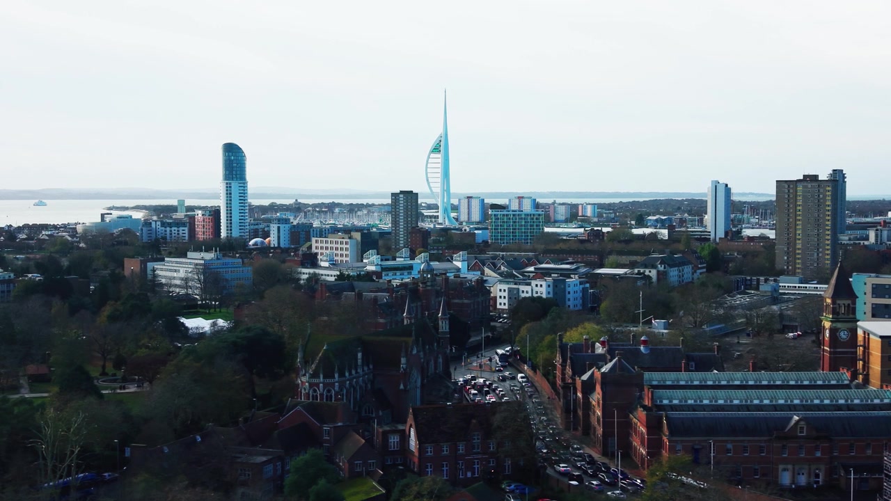 Riprese aeree con drone di Portsmouth che mostrano un campanile di chiesa, traffico intenso e tetti della città, con la Spinnaker Tower e il porto sullo sfondo.