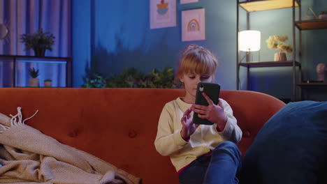 child toddler hold smartphone watching funny cartoons, chatting with friends on couch alone at home