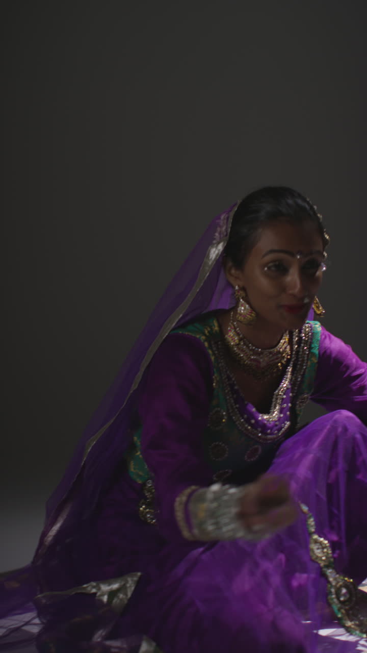Vertical Video Of Female Kathak Dancer Performing Dance Wearing Traditional Indian Dress Seated On Floor In Spotlight