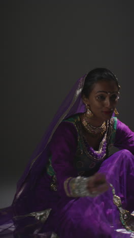 vertical video of female kathak dancer performing dance wearing traditional indian dress seated on floor in spotlight