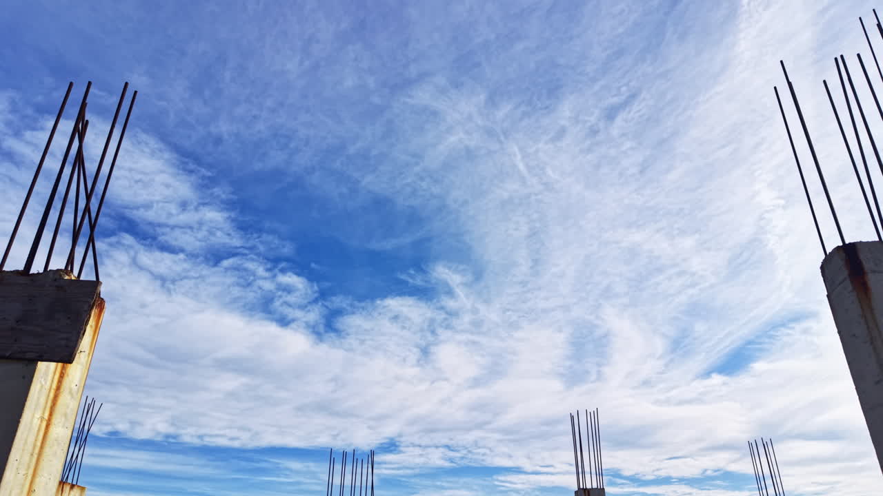 Pemandangan lokasi konstruksi ke arah langit dengan tiang-tiang beton yang menjulang ke awan di bawah langit biru cerah.