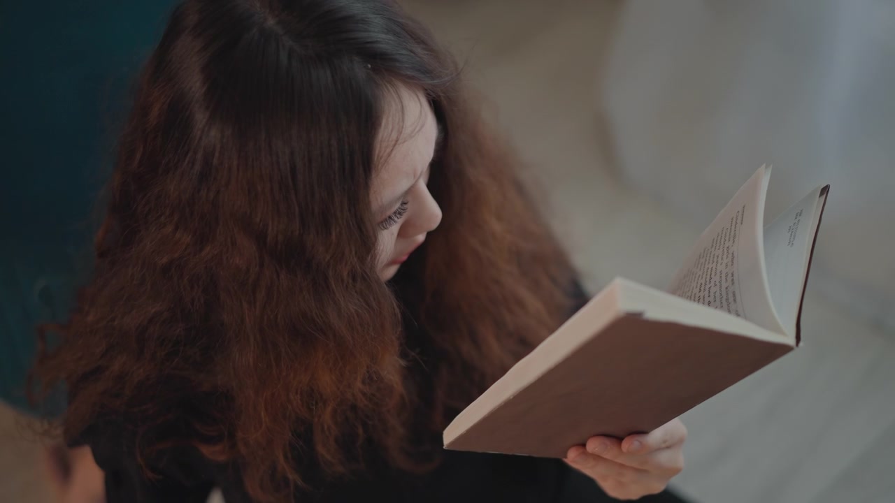 Giovane donna con lunghi capelli ricci castani tiene un libro aperto nella mano destra mentre le pagine si sollevano delicatamente, suggerendo una brezza e un movimento naturale e morbido durante una lettura tranquilla in un ambiente interno accogliente.