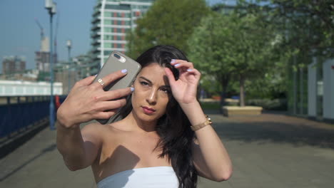 Beautiful-brunette-hispanic-model-woman-looks-at-her-phone-and-takes-selfies-next-to-the-river-Thames-in-London-during-bright-daylight-of-the-summer,-in-United-Kingdom