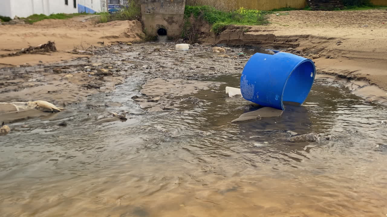 Dirty water from a drainage pipe flowing onto a sandy bank with a blue plastic barrel and other garbage, showing pollution