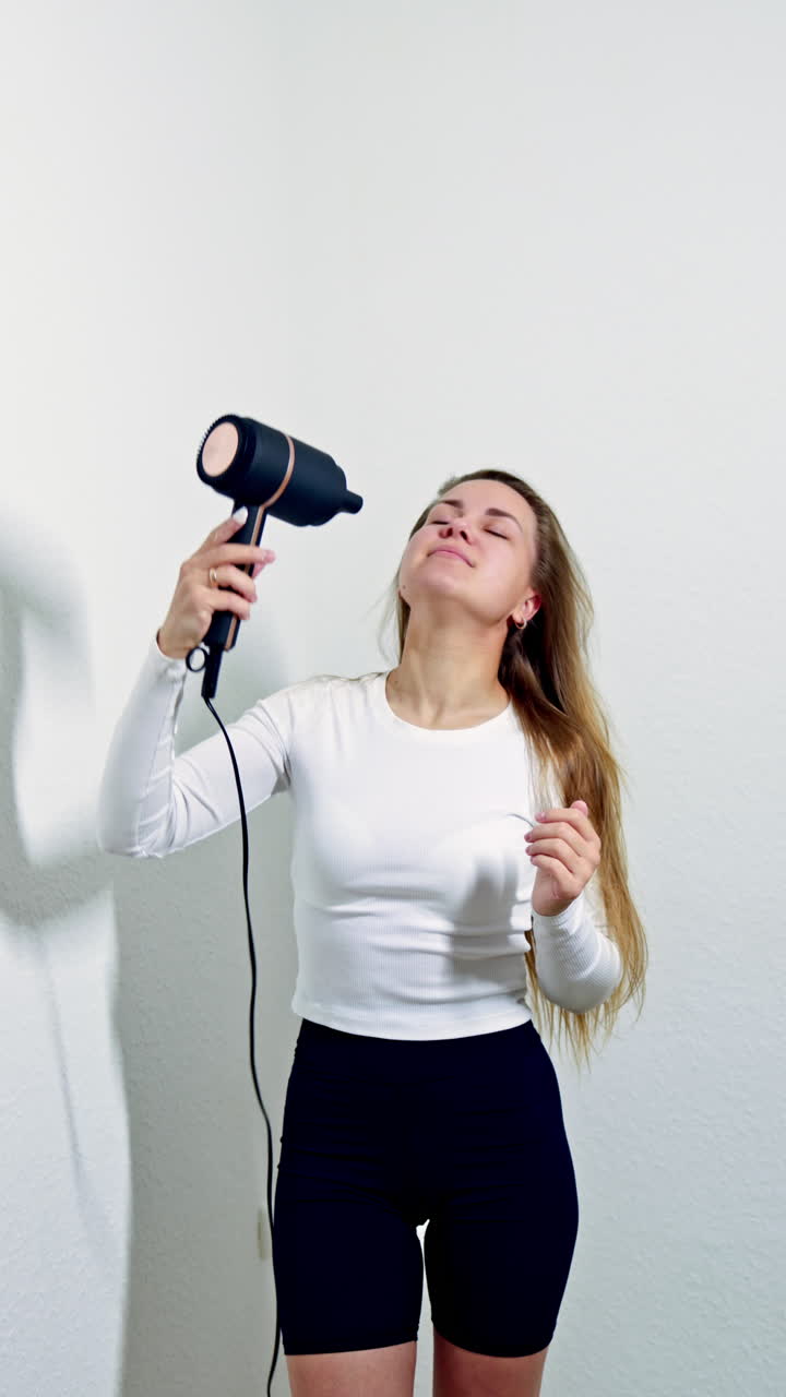 Una mujer hermosa, después de lavarse el cabello, se lo seca con un secador y canta. Una joven admira su cabello tras los procedimientos de cuidado. Cuidado para piel dorada y cabello largo.