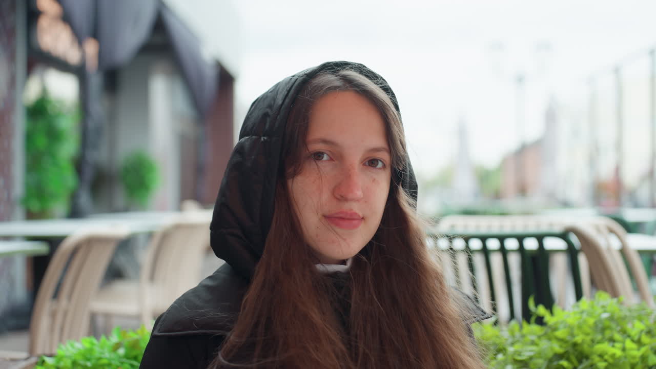 Giovane donna bianca seduta in un bar, con felpa con cappuccio e maglione, lunghi capelli che le incorniciano il viso, occhi chiusi che poi si aprono, luce soffusa e diffusa, sfondo sfocato con strada di città, fioriere verdi e panchina di metallo