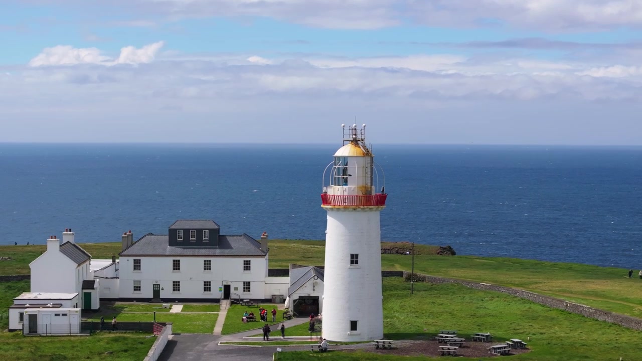 Un faro bianco si erge imponente sullo sfondo dell'oceano blu, circondato da edifici e campi verdi. Adatto per temi di viaggio e marittimi.