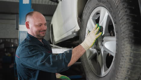 mechanic in blue uniform with hairy hand and white gloves smiling while adjusting and aligning car tire in auto repair workshop, ensuring proper positioning and safety