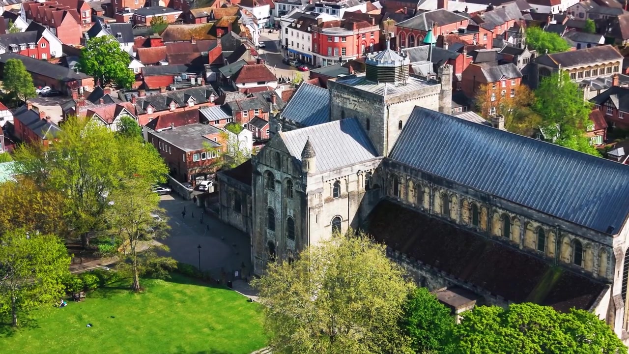 Riprese aeree ravvicinate con drone dell'Abbazia di Romsey nell'Hampshire, che mostrano la storica chiesa abbaziale, l'architettura in pietra, i dettagli del tetto, le strade circostanti, gli edifici della città, gli alberi e gli spazi verdi in una giornata di sole