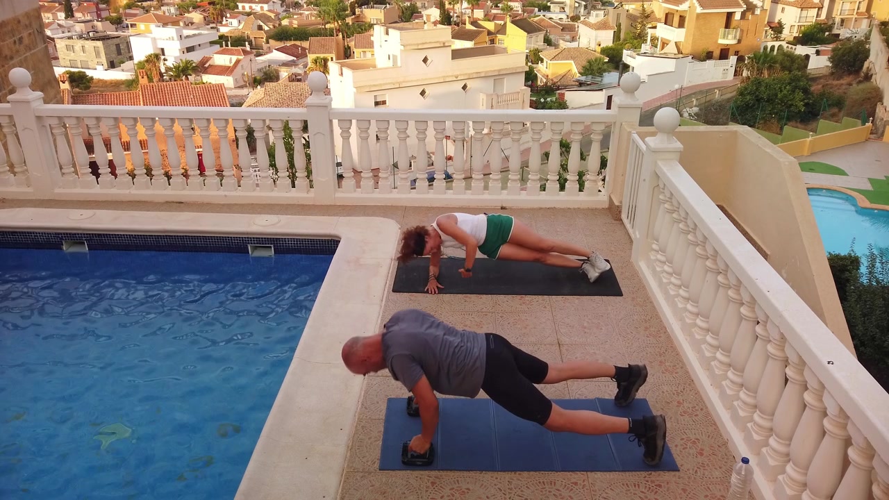High angle shot of a man using push up bars and a woman performing side plank arm reaches on exercise mats beside a luxury villa swimming pool.