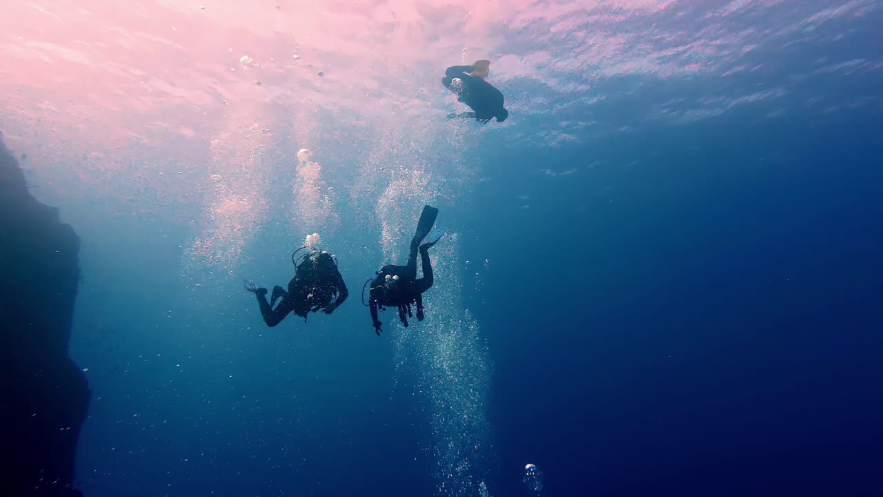 Three adventurous scuba divers explore deep crystalline blue oceanic waters near a jagged dark underwater cliff during a bright summer day featuring rising air bubbles and cinematic wide low angles