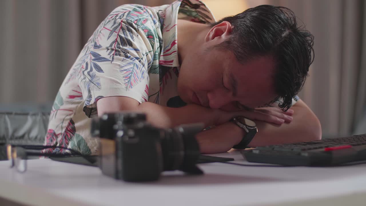 체어에 앉아 있는 피곤한 아시아인 남자가 집에 있는 카메라 에 있는 데스크에서 비디오를 편집하는 동안 잠을 자고 있다.