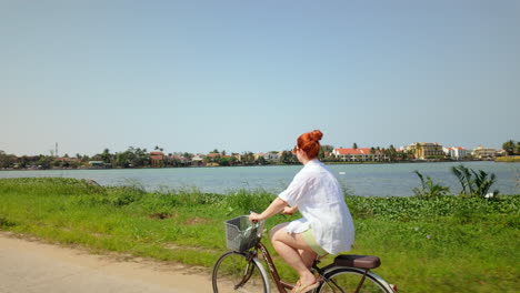 woman riding bicycle along the river on a sunny day in hoi an, vietnam. - wide shot