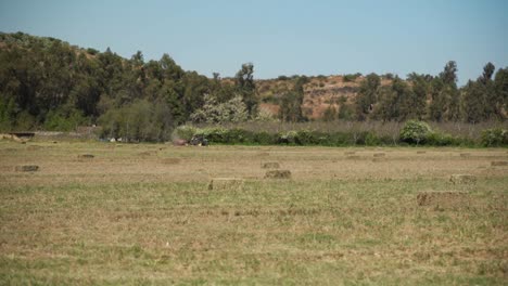 Strohballen,-Die-Von-Einem-Traktor-Im-Hintergrund-Verdichtet-Werden,-Flacher-Hintergrund-Mit-Einem-Berg-Im-Hintergrund