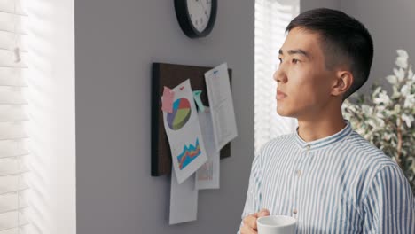 serious focused thoughtful head of the company, the businessman stands in the office of the corporate at the window covered with blinds and looks into it, contemplates the business