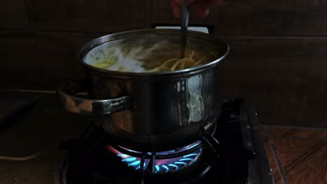 Spaghetti-being-stirred-in-a-silver-pot-that-stands-on-a-gas-stove-in-front-of-dark-background