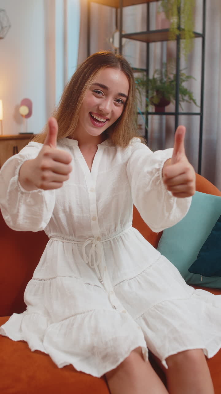 Portrait of excited smiling young woman showing double thumbs up sitting on sofa couch at home