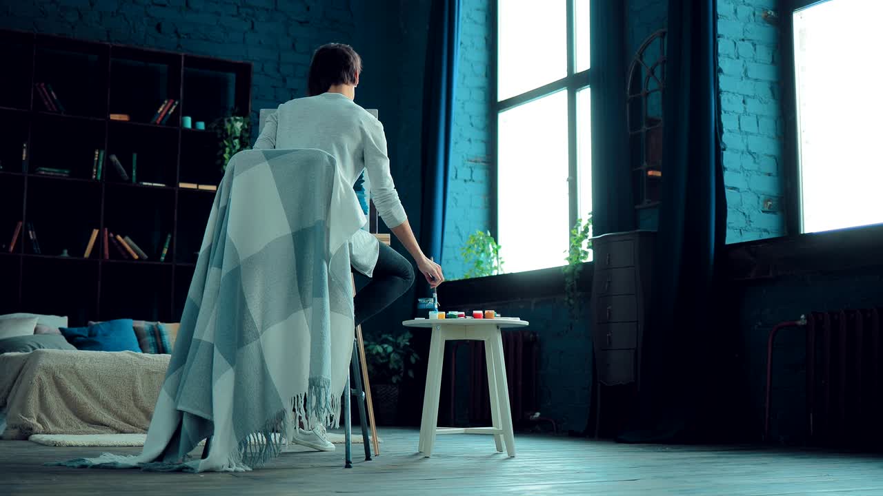 젊은 소녀는 종이에 그림을 그립니다. 시트는 캔버스에 서 있습니다, 테이블에 그림을 칠합니다. 가운 방. 카메라의 원형 움직임.