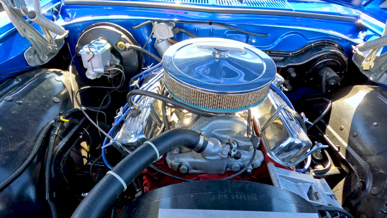 A detailed close-up pan of a retro car engine bay in bright daylight, highlighting chrome components and blue bodywork at an outdoor event