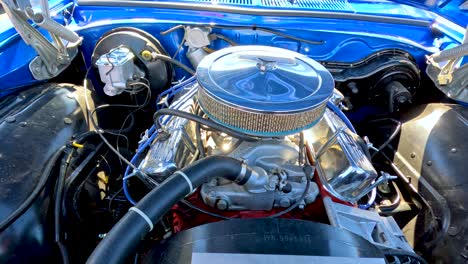 a detailed close-up pan of a retro car engine bay in bright daylight, highlighting chrome components and blue bodywork at an outdoor event