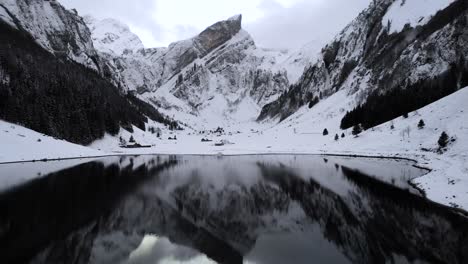 Aerial-flyover-across-lake-Seealpsee-in-Appenzell,-Switzerland-surrounded-by-snow-on-a-winter-day-with-a-reflection-of-the-Alpstein-peaks-on-the-calm-water