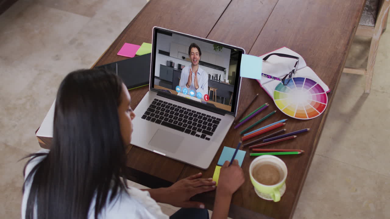 mujer caucásica usando una computadora portátil en videollamada con un colega masculino, tomando notas