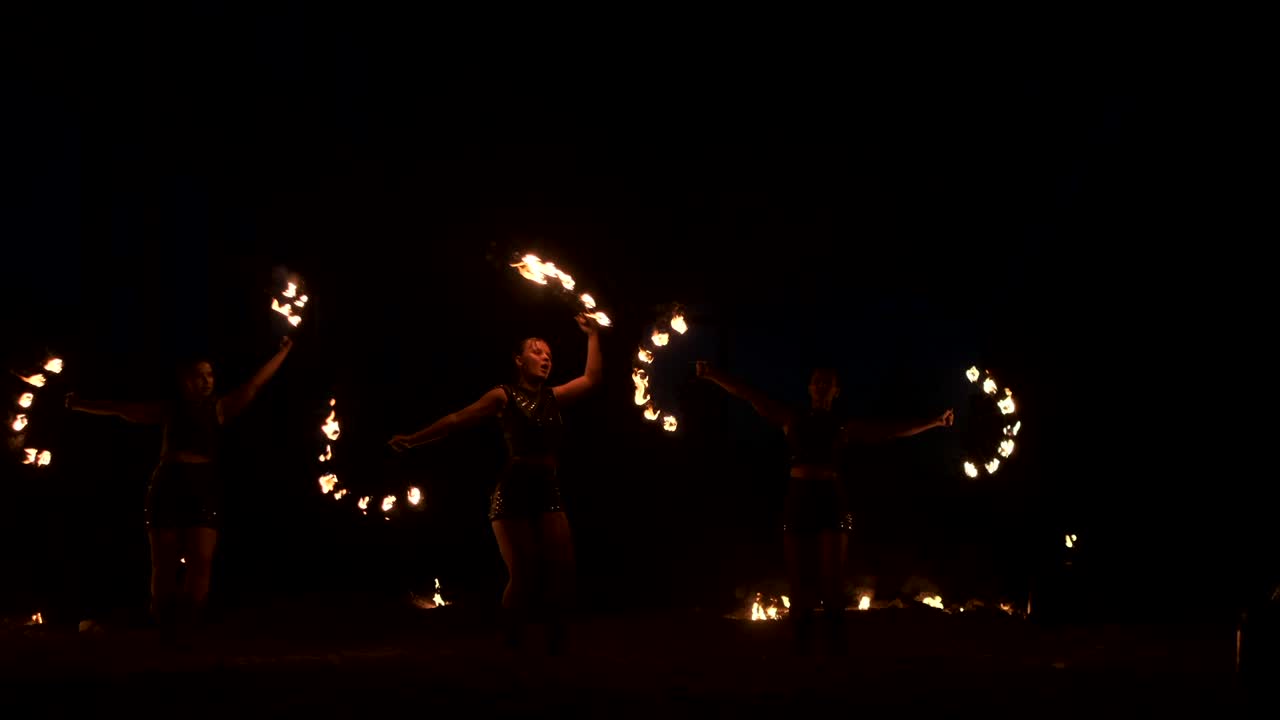 tres mujeres con aros ardientes bailan con antorchas ardientes en ropa de cuero en un hangar oscuro demostrando un espectáculo de fuego de circo en cámara lenta.