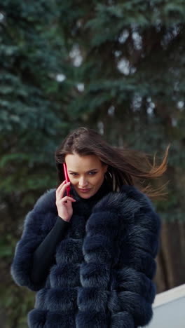 luxury girl with a phone in the park. portrait of attractive lady in fur coat walking in the park and talking the mobile phone. slow motion. vertical video