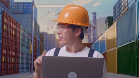 close up de un trabajador asiático que usa gafas de protección y casco de seguridad usando una computadora portátil y apuntando hacia un lado mientras está de pie en un almacén de contenedores