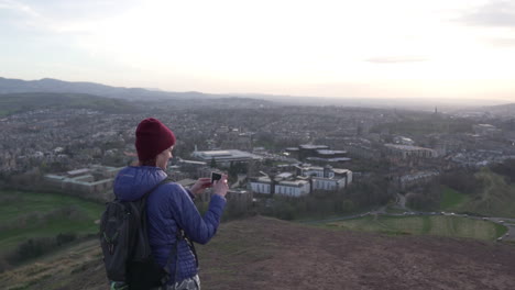 Kreisende-Parallaxen-Zeitlupenaufnahme-Eines-Mädchens,-Das-Am-Abend-Ein-Foto-Mit-Dem-Smartphone-Vom-Arthurs-Seat-Mountain-Macht,-Mit-Der-Stadt-Edinburgh-Im-Hintergrund-Während-Einer-Wundervollen-Goldenen-Stunde-Mit-Sonnenuntergang