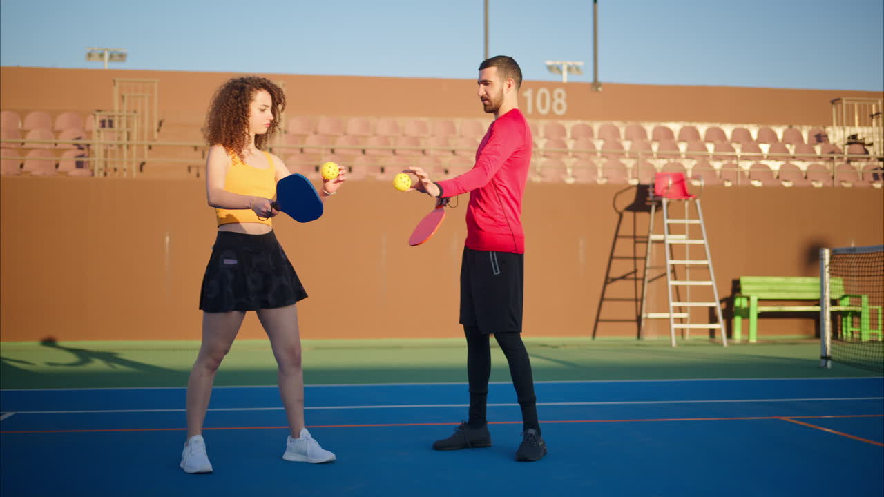 A man in a red shirt teaching a woman with curly hair how to play pickleball on a blue court on a sunny day